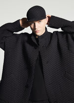 Model wears TYPE-O 024 by A-POC ABLE ISSEY MIYAKE, a textured black coat in double-weave fabric with a matching cap, adjusting the hat while looking at the camera against a plain light background.