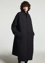 A person with curly hair wears the TYPE-O 024 long, textured black coat by A-POC ABLE ISSEY MIYAKE, hands in pockets, standing against a plain light background.