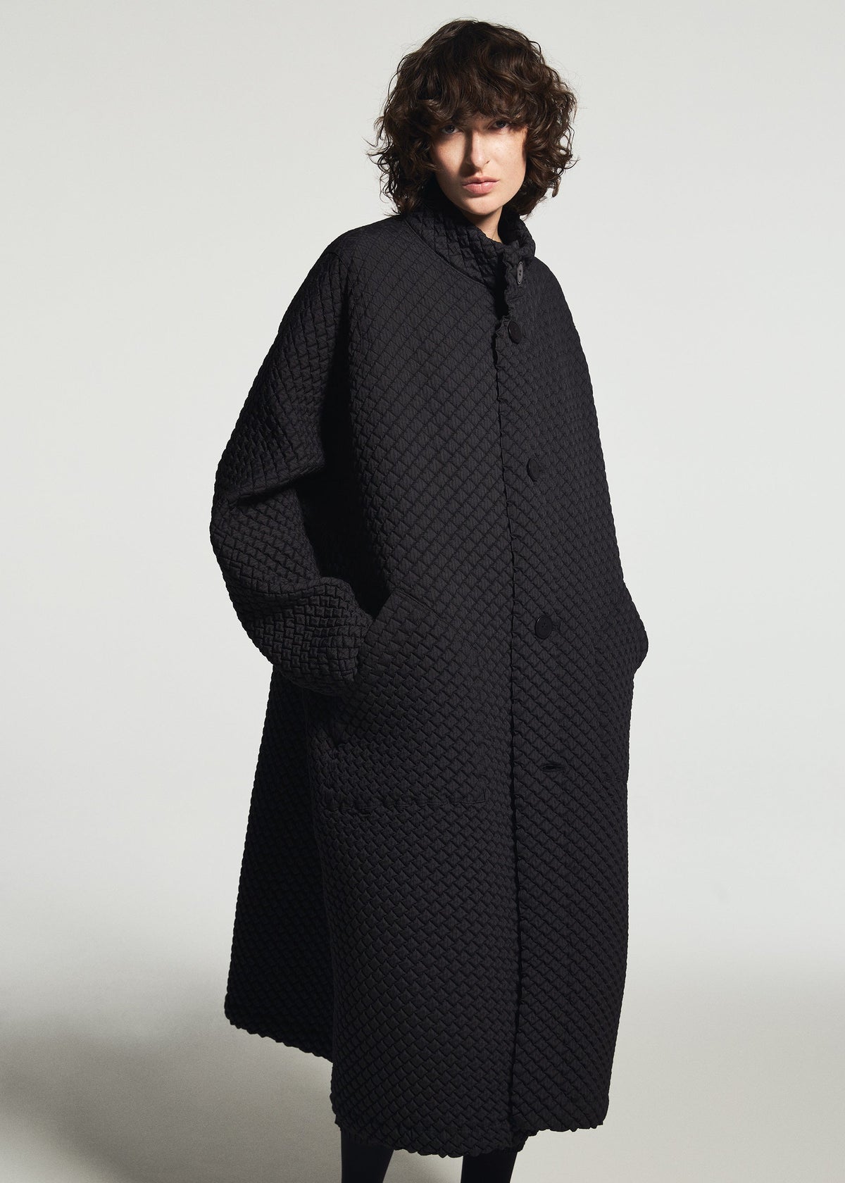 Zoomed view A person with curly hair wears the TYPE-O 024 long, textured black coat by A-POC ABLE ISSEY MIYAKE, hands in pockets, standing against a plain light background.