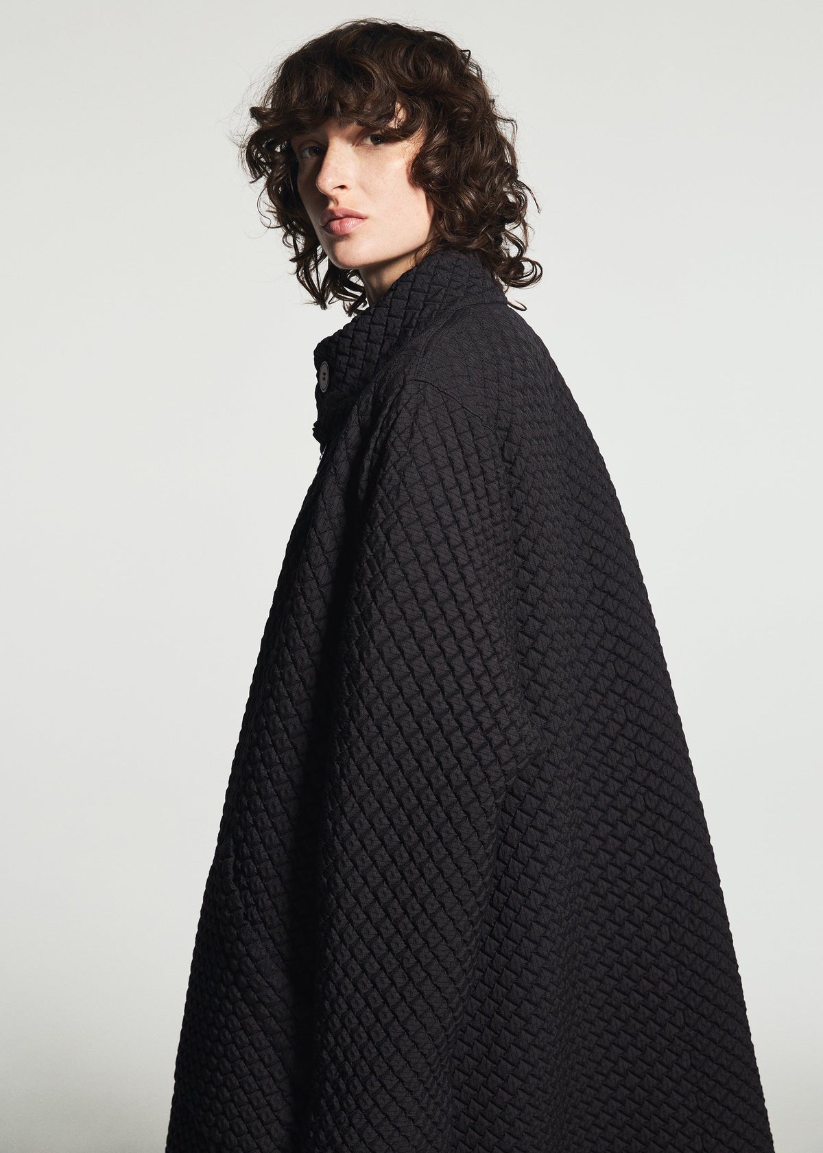 A person with shoulder-length curly hair wears the TYPE-O 024 oversized double-weave coat by A-POC ABLE ISSEY MIYAKE, standing against a light background and looking over their shoulder with a neutral expression. view 13 of 15