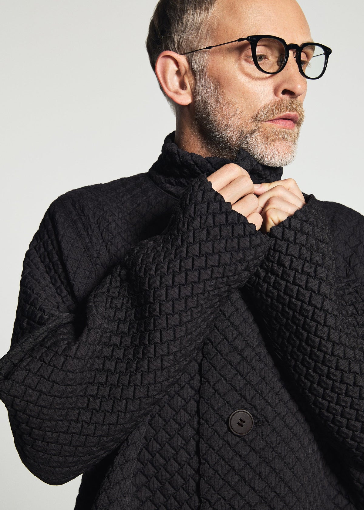 Zoomed view A man with glasses and a gray beard wears the TYPE-O 024 jacket by A-POC ABLE ISSEY MIYAKE, holding up the collar with both hands and gazing slightly to the side against a light background.