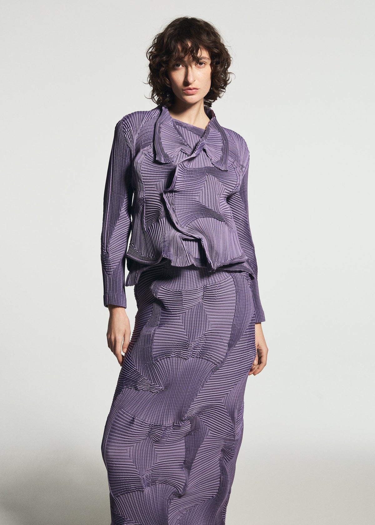Zoomed view A person with short curly hair wears the TYPE-O 003-1 textured, purple long-sleeve top and matching skirt by A-POC ABLE ISSEY MIYAKE, featuring abstract geometric patterns crafted from unique blends of recycled polyester, standing against a plain light background.