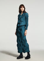 A woman stands against a plain white background wearing the TYPE-O 003-1 set by A-POC ABLE ISSEY MIYAKE—a textured teal long-sleeve blouse and matching ankle-length skirt—paired with black boots.