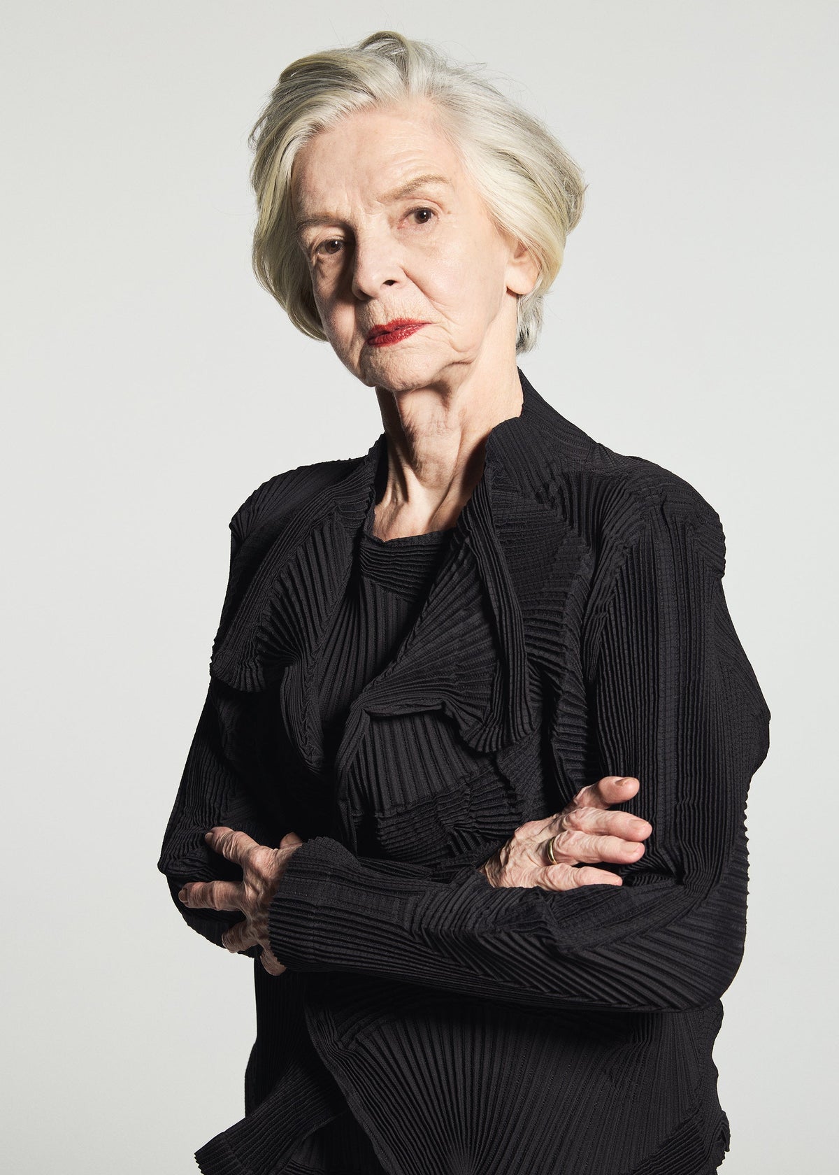 A woman with short gray hair stands with her arms crossed and a serious expression against a plain white background, wearing the TYPE-O 003 BK by A-POC ABLE ISSEY MIYAKE—a pleated black outfit made from recycled polyester and distinguished by its unique textile design. view 3 of 3