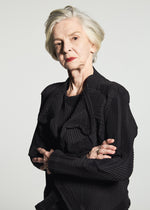 A woman with short gray hair stands with her arms crossed and a serious expression against a plain white background, wearing the TYPE-O 003 BK by A-POC ABLE ISSEY MIYAKE—a pleated black outfit made from recycled polyester and distinguished by its unique textile design.