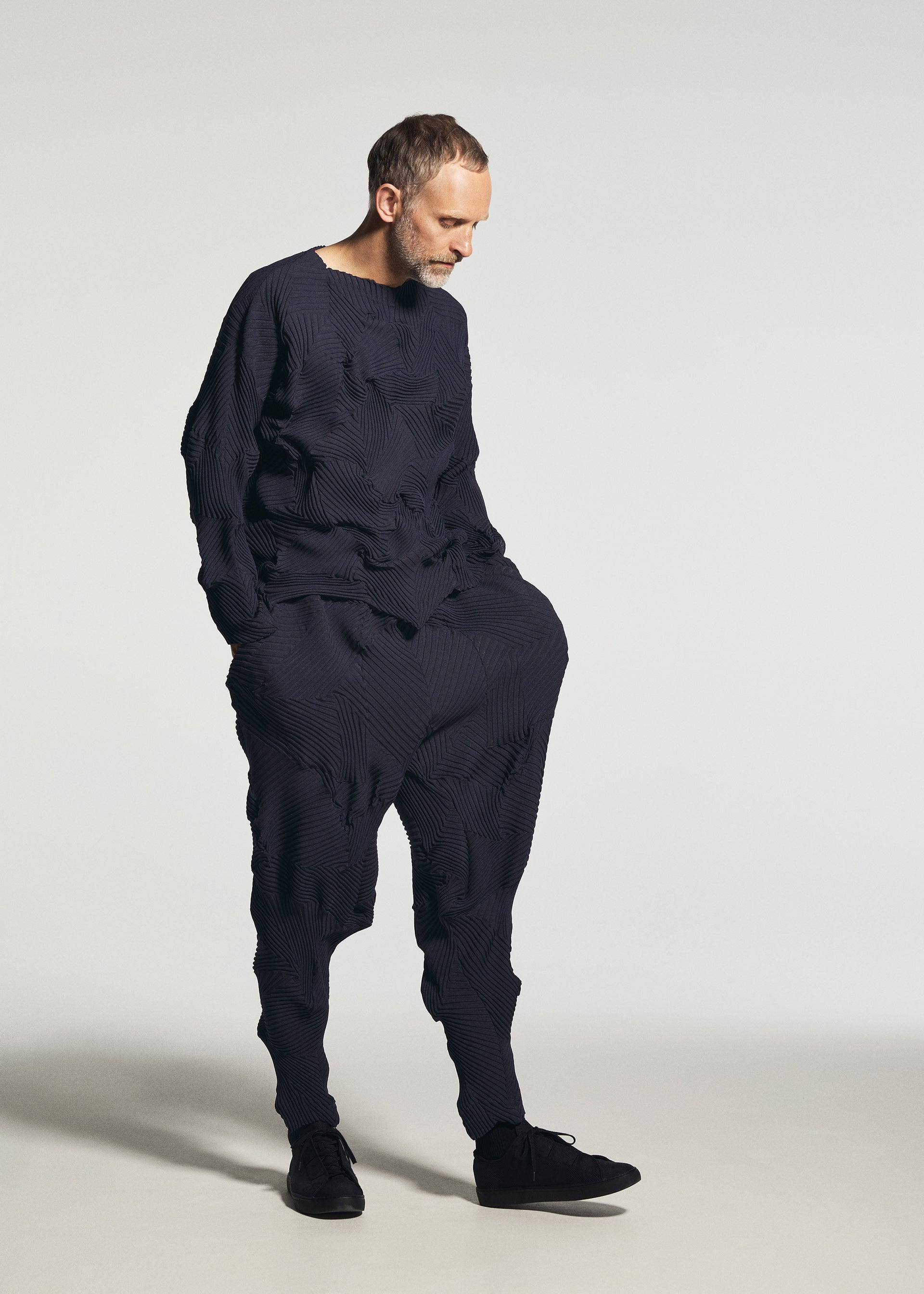 Zoomed view Wearing the TYPE-P 003-1 by A-POC ABLE ISSEY MIYAKE, a man with short hair and a beard stands against a plain light background. He looks down, dressed in a loose-fitting, textured navy blue ensemble featuring pleated pants and black shoes.