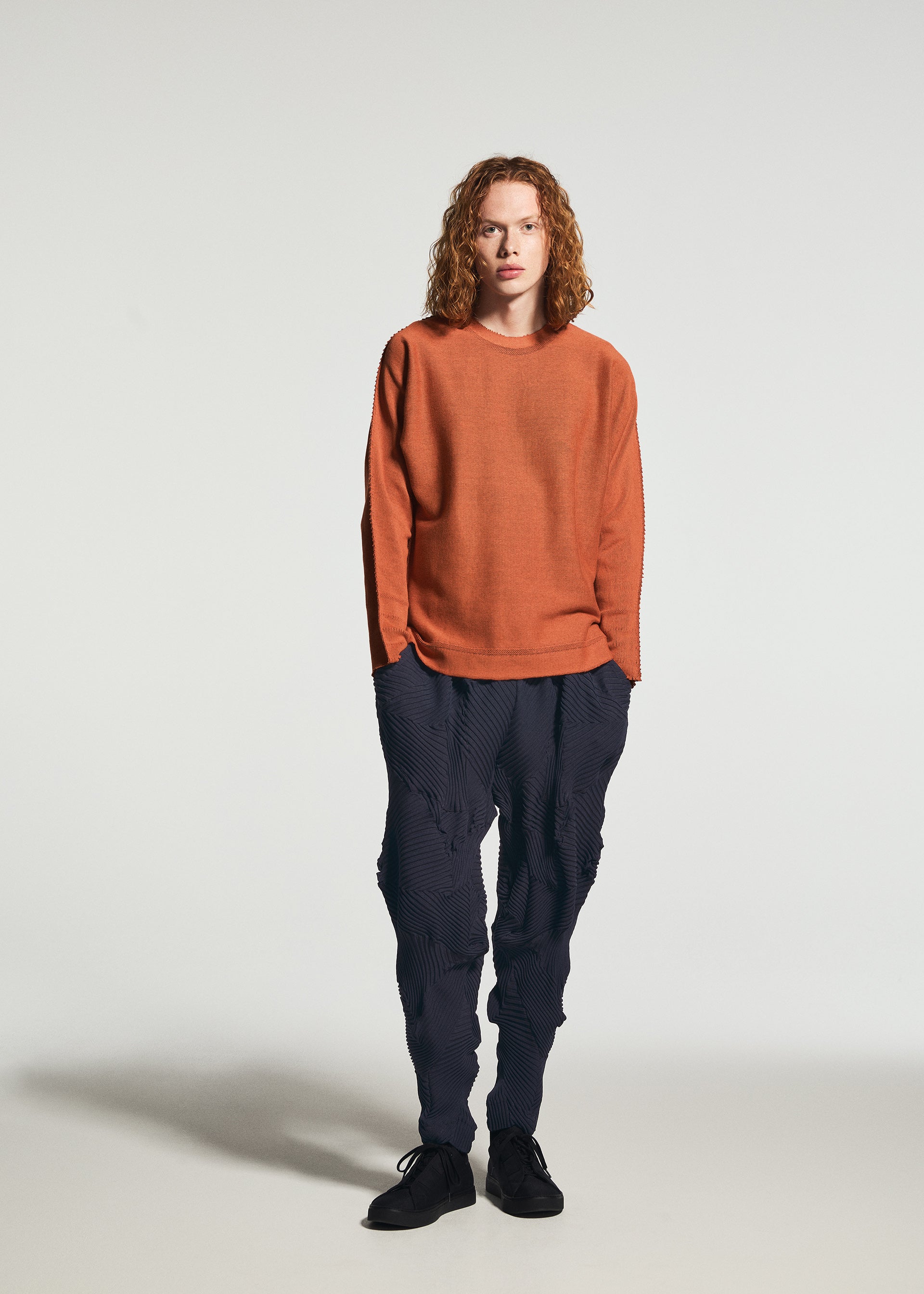 Zoomed view A person with curly hair stands against a plain background, wearing TYPE-P 003-1 by A-POC ABLE ISSEY MIYAKE—an orange long-sleeve shirt paired with textured pleated pants and black shoes, showcasing a look perfect for versatile everyday wear.