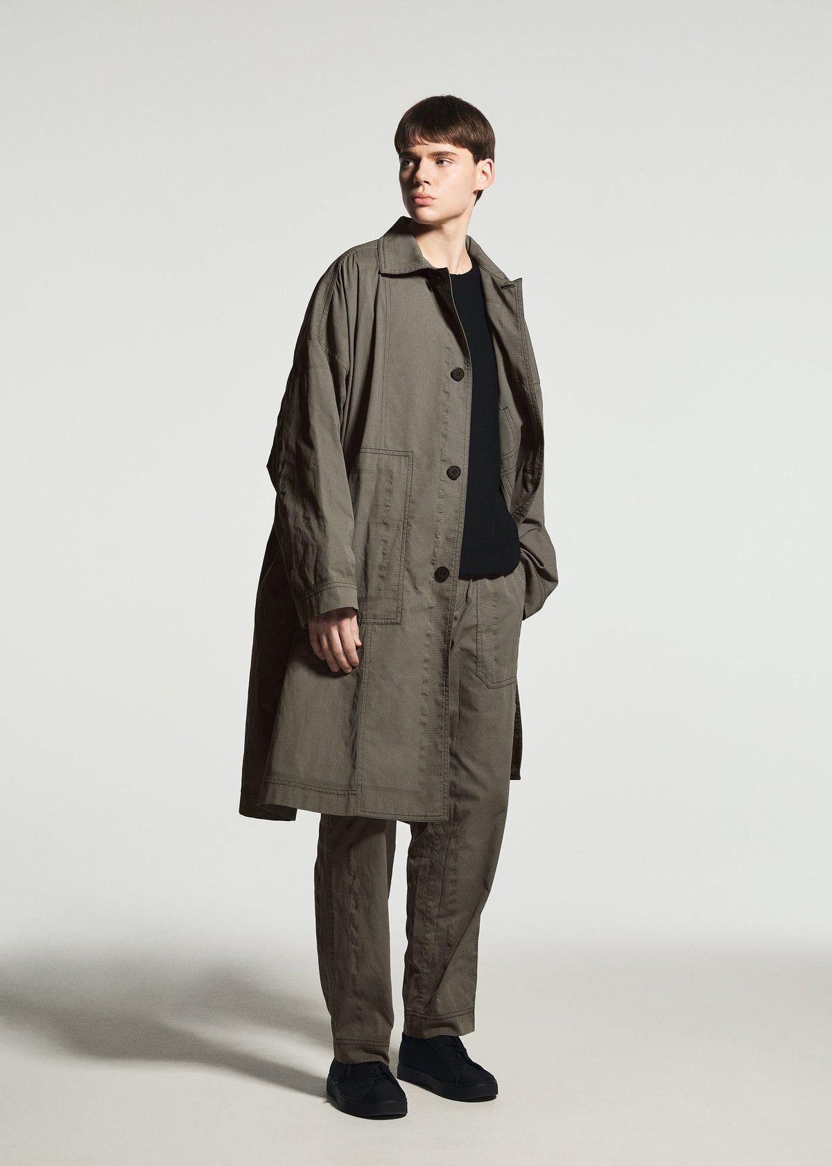 Standing against a plain light background, the person wears the TYPE-U 001-1 by A-POC ABLE ISSEY MIYAKE—a loose-fitting khaki trench coat layered over coordinating wrinkle resistant pants and a black shirt, paired with black shoes. view 5 of 6