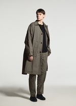 Standing against a plain light background, the person wears the TYPE-U 001-1 by A-POC ABLE ISSEY MIYAKE—a loose-fitting khaki trench coat layered over coordinating wrinkle resistant pants and a black shirt, paired with black shoes.
