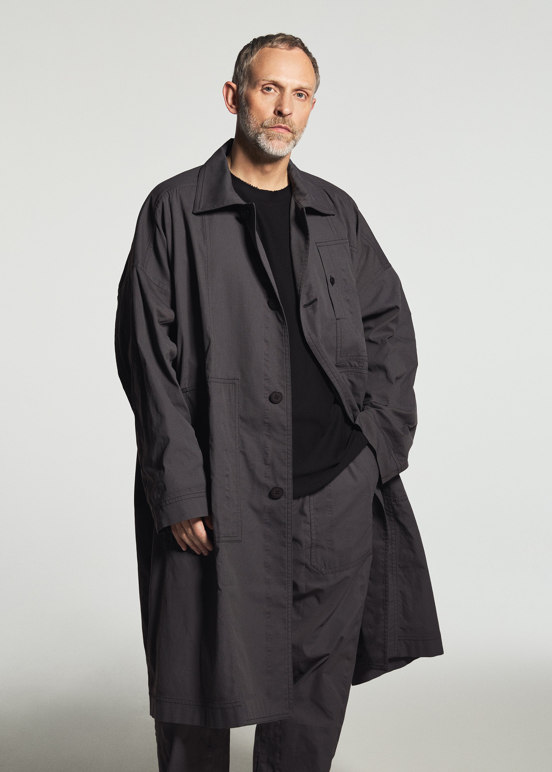 Zoomed view A man with short hair and a beard stands against a plain, light gray background, wearing the TYPE-U 001-1 by A-POC ABLE ISSEY MIYAKE—a long, dark trench coat layered over a black ensemble featuring adjustable straight pants.