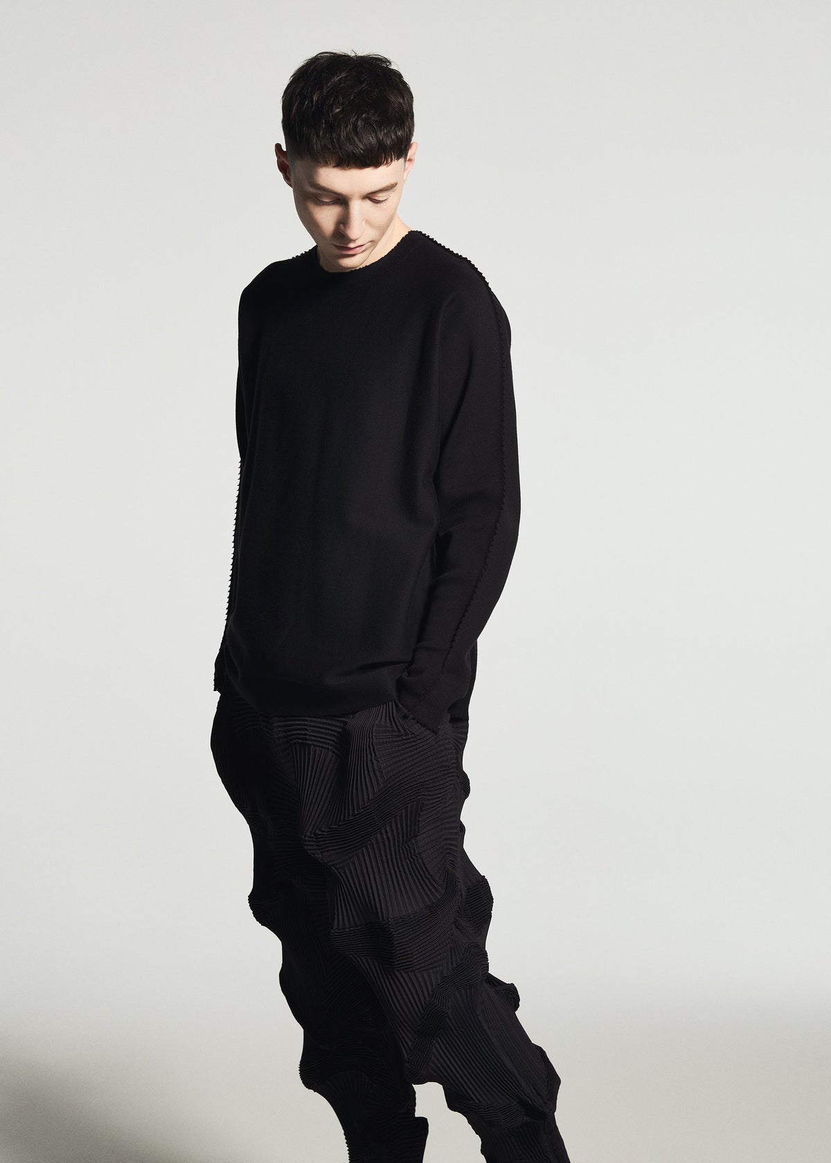 Wearing the TYPE-O 003 BK textured black Steam Stretch pants crafted from recycled polyester by A-POC ABLE ISSEY MIYAKE and a black long-sleeve shirt, a person stands against a plain white background, looking down with hands in pockets. view 5 of 7