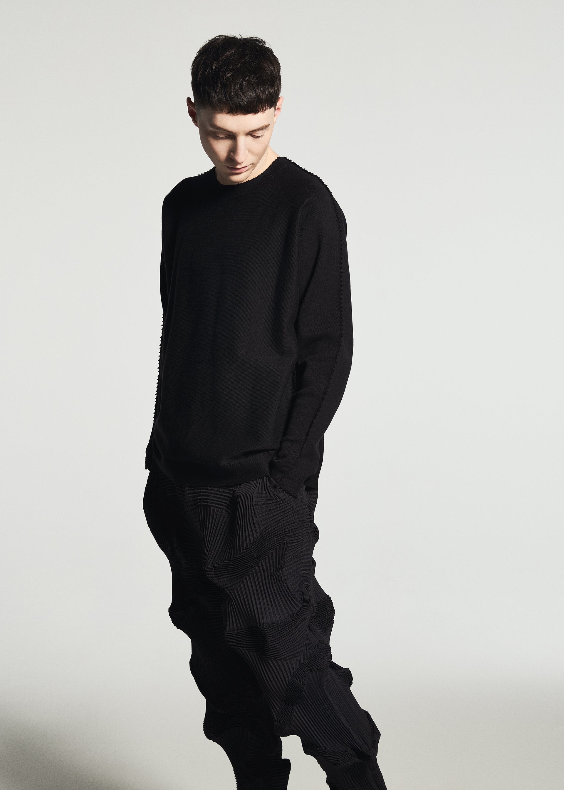 Zoomed view Wearing the TYPE-O 003 BK textured black Steam Stretch pants crafted from recycled polyester by A-POC ABLE ISSEY MIYAKE and a black long-sleeve shirt, a person stands against a plain white background, looking down with hands in pockets.