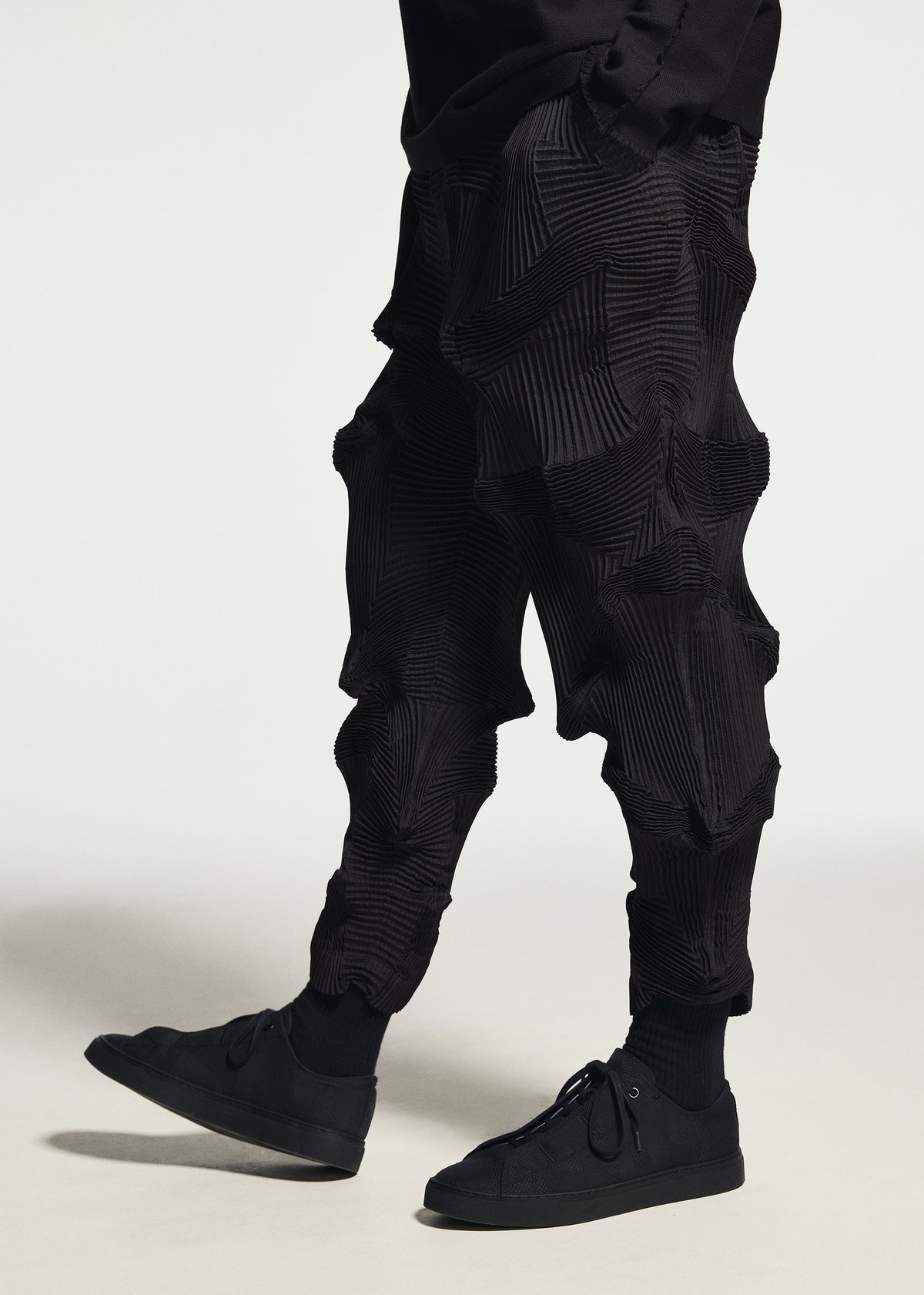 A person wearing TYPE-O 003 BK textured, pleated black Steam Stretch pants by A-POC ABLE ISSEY MIYAKE—crafted from a unique recycled polyester textile with sculptural shapes—paired with black sneakers and standing against a plain white background. view 6 of 7