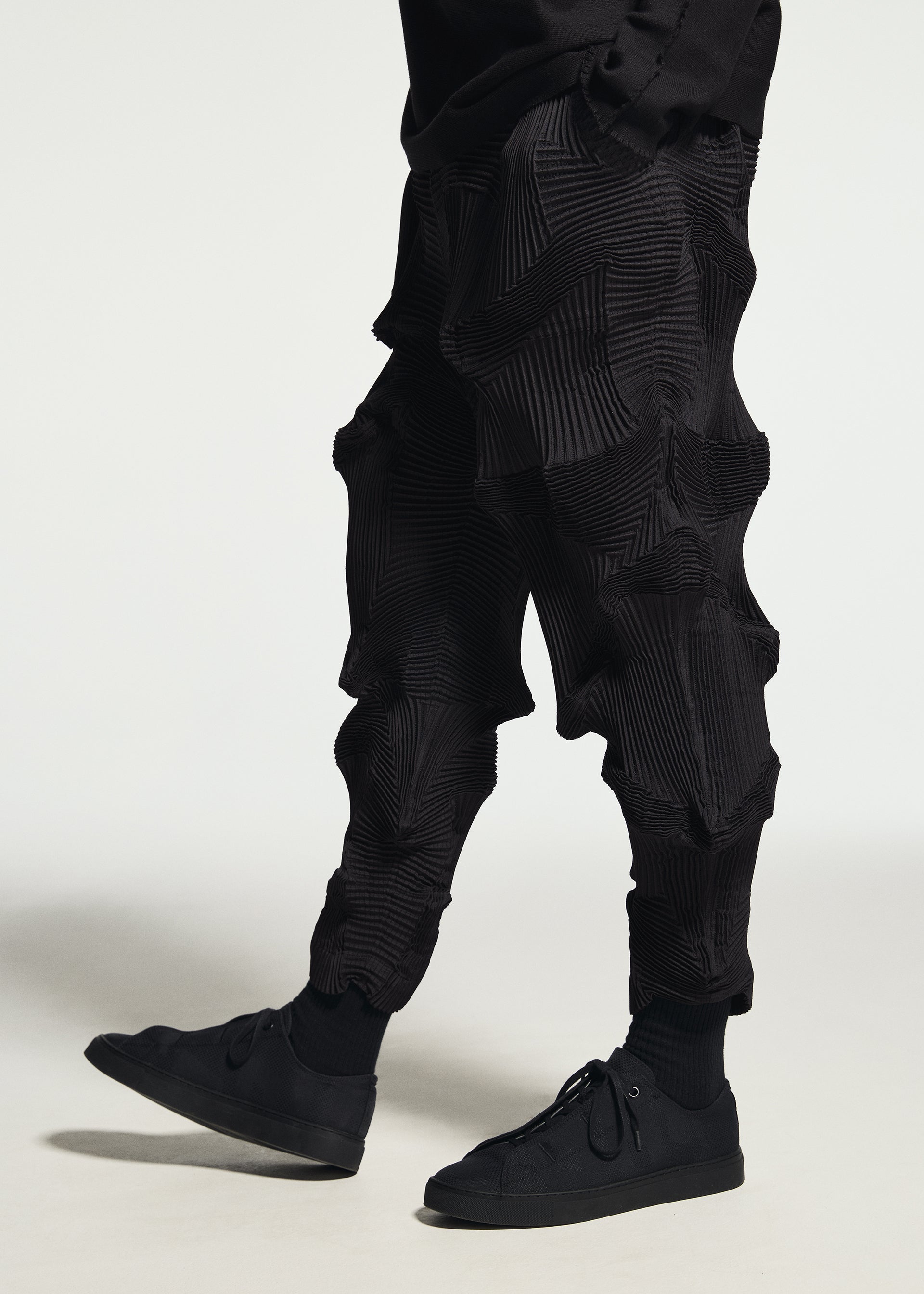 Zoomed view A person wearing TYPE-O 003 BK textured, pleated black Steam Stretch pants by A-POC ABLE ISSEY MIYAKE—crafted from a unique recycled polyester textile with sculptural shapes—paired with black sneakers and standing against a plain white background.