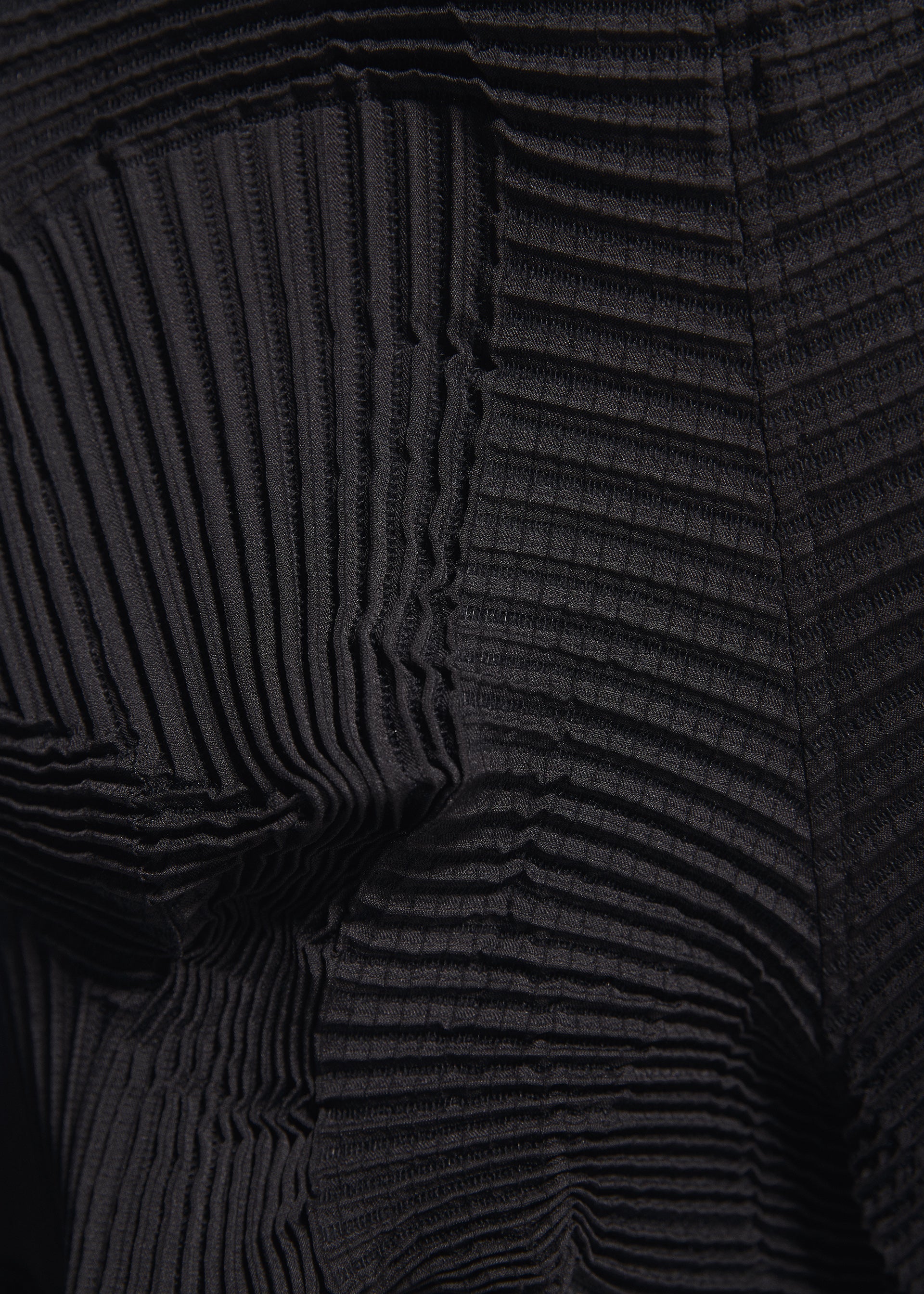 Zoomed view Close-up of black pleated fabric with textured, wavy ridges and geometric folds, showcasing varying depths and shadows. Crafted from recycled polyester, this unique textile is featured in the TYPE-O 003 BK pants by A-POC ABLE ISSEY MIYAKE.