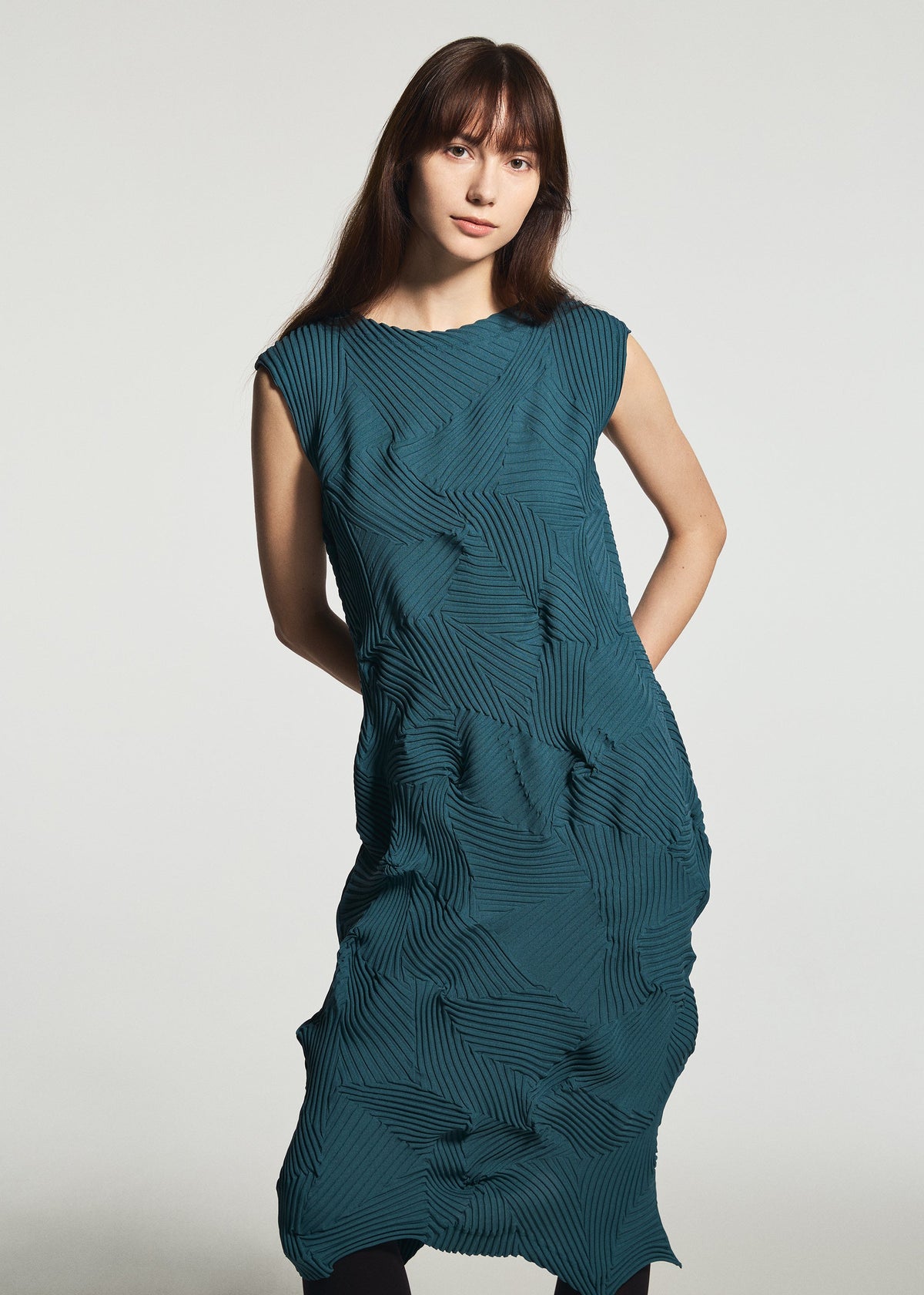 A woman with long brown hair wears the TYPE-P 003-1 sleeveless teal dress by A-POC ABLE ISSEY MIYAKE, featuring a textured geometric pattern. Standing against a plain light background, she showcases the effortless style and comfort of this versatile everyday piece. view 5 of 6