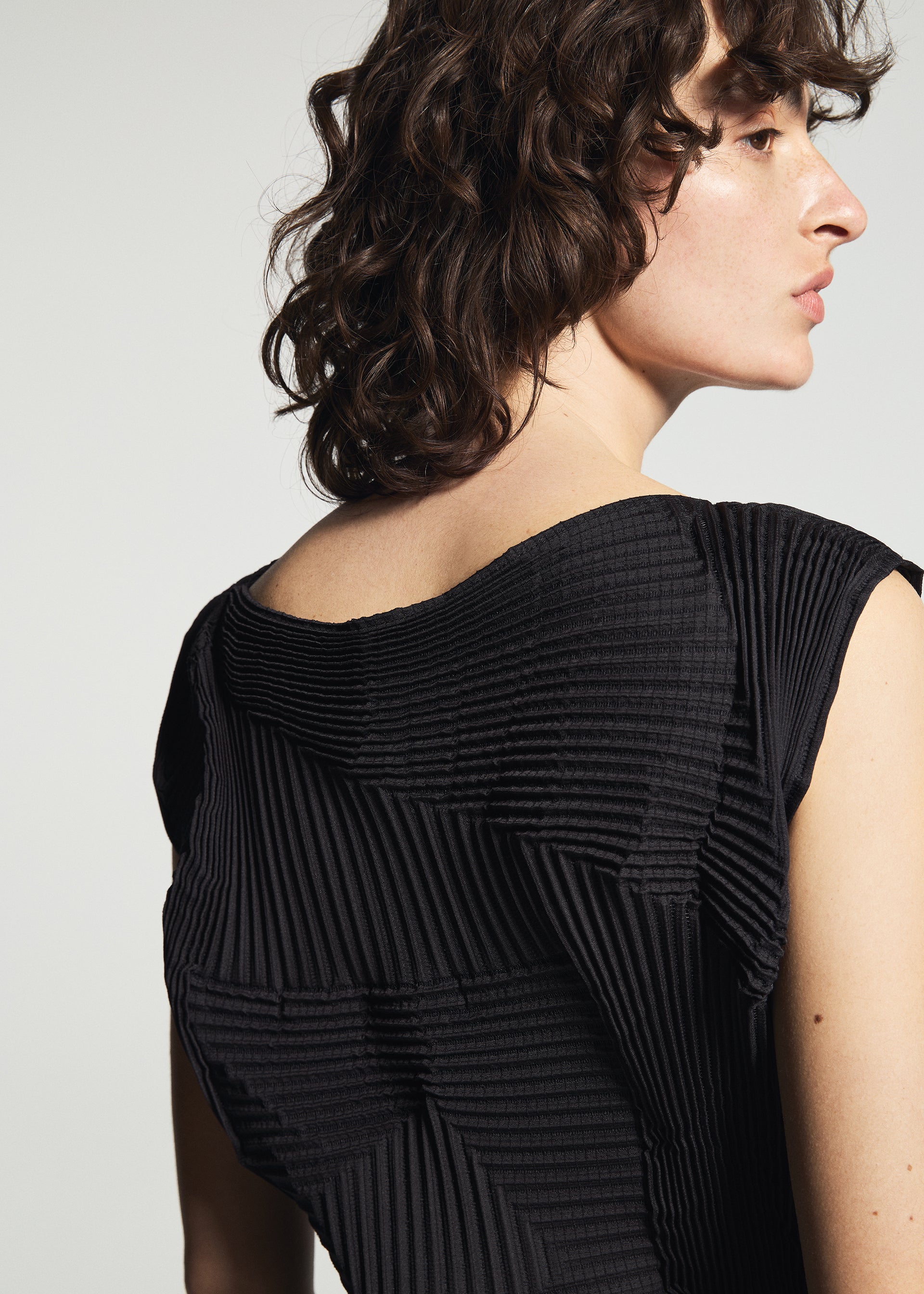 Zoomed view Wearing the TYPE-O 003 BK textured black top by A-POC ABLE ISSEY MIYAKE, a person with wavy brown hair stands sideways, looking away from the camera; the geometric ribbed patterns are inspired by Baked Stretch.