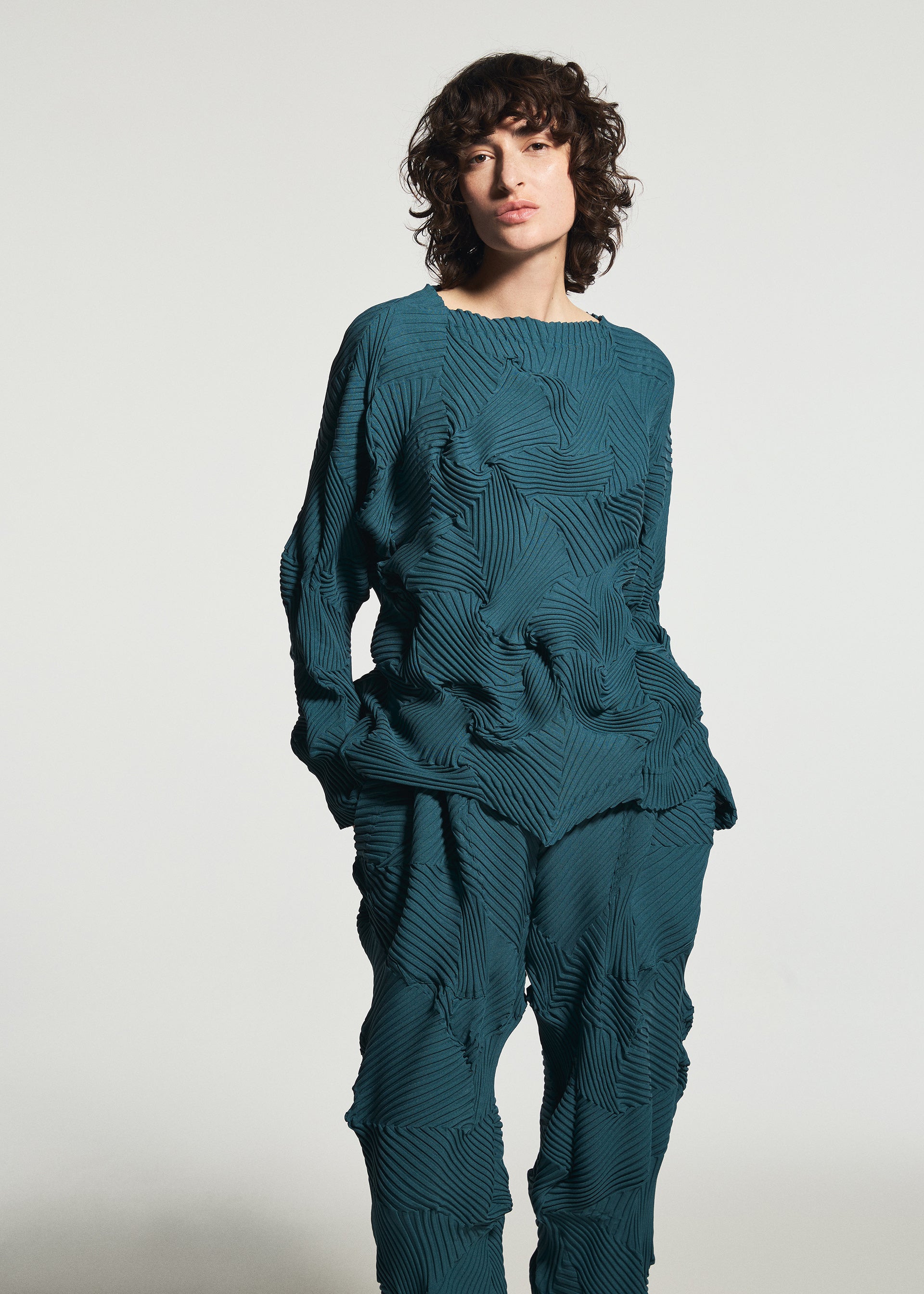 Zoomed view Wearing the TYPE-P 003-1 set from A-POC ABLE ISSEY MIYAKE, a person with curly hair poses against a plain light gray background in the teal, long-sleeved textured top and matching geometric-patterned pants.