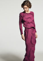 A person wearing the A-POC ABLE ISSEY MIYAKE TYPE-O 003-1 dress—a long-sleeved, textured magenta garment with geometric patterns crafted from recycled polyester—looks down while standing against a plain background.