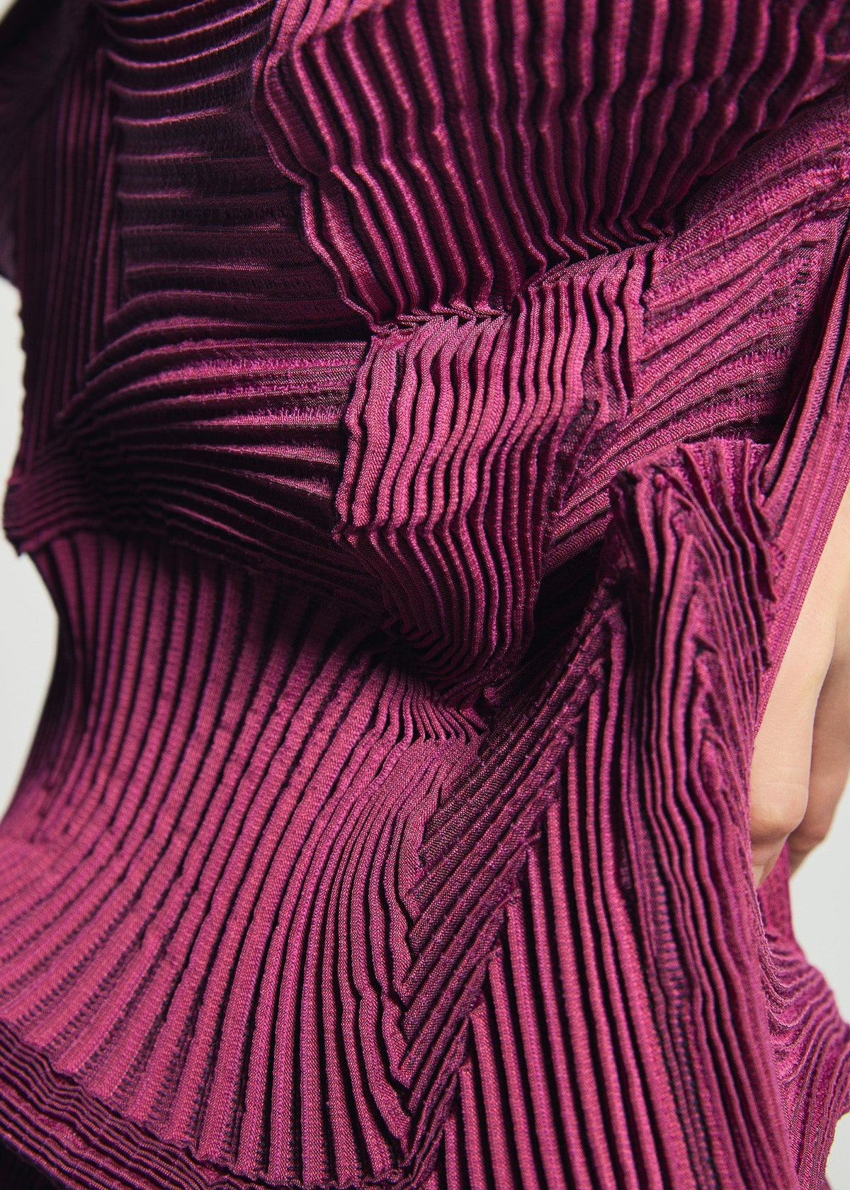 Zoomed view Close-up of the TYPE-O 003-1 top by A-POC ABLE ISSEY MIYAKE in magenta, crafted from recycled polyester and distinguished by its intricate three-dimensional pleats and geometric folds that form a textured, architectural pattern. Part of a person’s hand and waist are partially visible.
