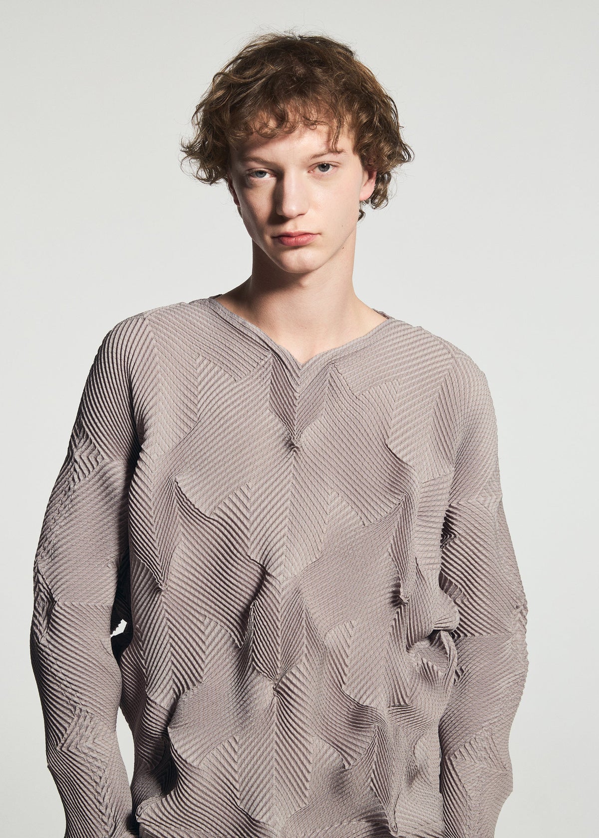 Zoomed view A young person with short, curly brown hair wears the TYPE-O 010-1 by A-POC ABLE ISSEY MIYAKE, a textured light mauve pleated long-sleeved top with a star pattern, standing against a plain light gray background.