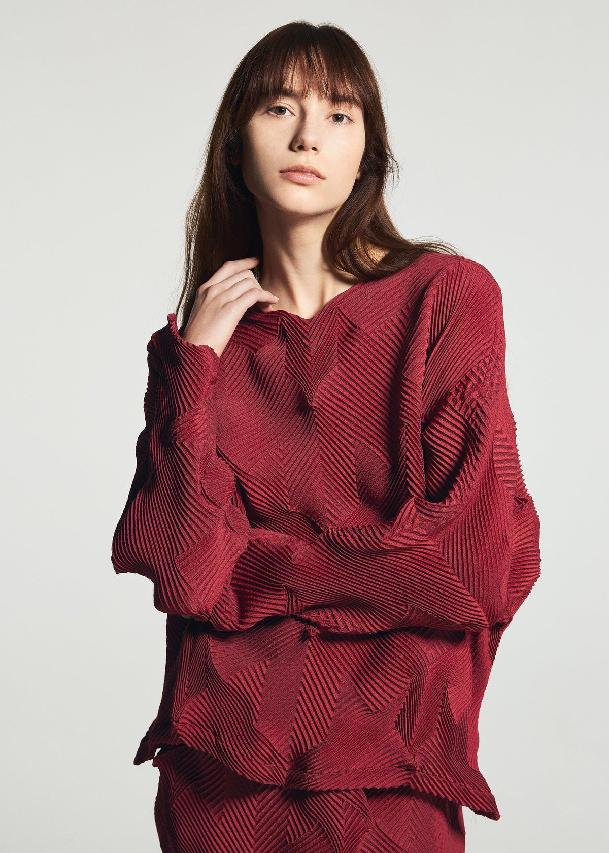 Zoomed view A person with long brown hair wears the TYPE-O 010-1 by A-POC ABLE ISSEY MIYAKE, a pleated dark red recycled polyester top, standing against a light background and looking at the camera with a neutral expression.