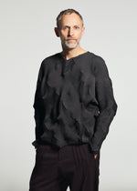 A man with short hair and a beard wears the TYPE-O 010-1 by A-POC ABLE ISSEY MIYAKE— a geometric-patterned, pleated long-sleeved top—paired with dark pants against a plain light background.