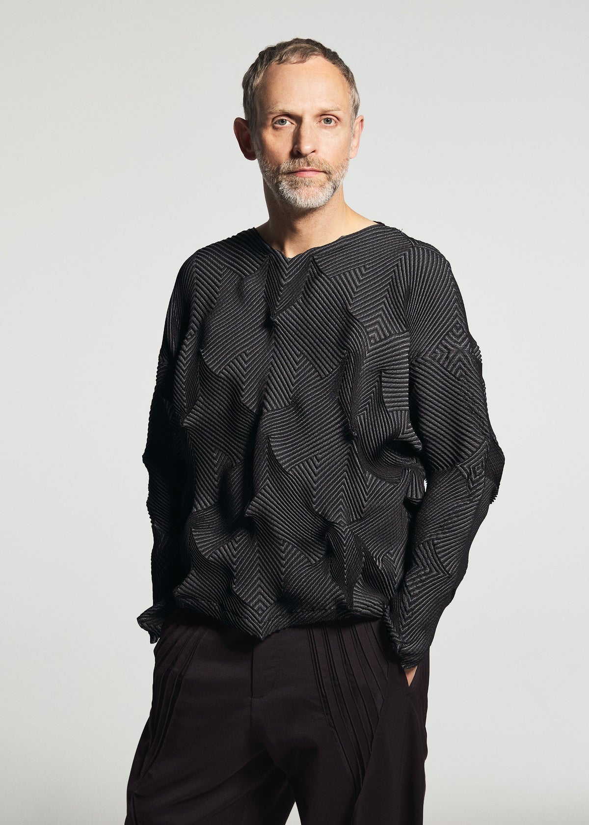Zoomed view A man with short hair and a beard wears the TYPE-O 010-1 by A-POC ABLE ISSEY MIYAKE— a geometric-patterned, pleated long-sleeved top—paired with dark pants against a plain light background.