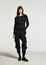 A woman stands against a plain background, wearing the TYPE-O 003 BK set from A-POC ABLE ISSEY MIYAKE—a textured black long-sleeve top and matching pants, both crafted from plant-derived polyester with wavy, sculpted details—and black shoes.