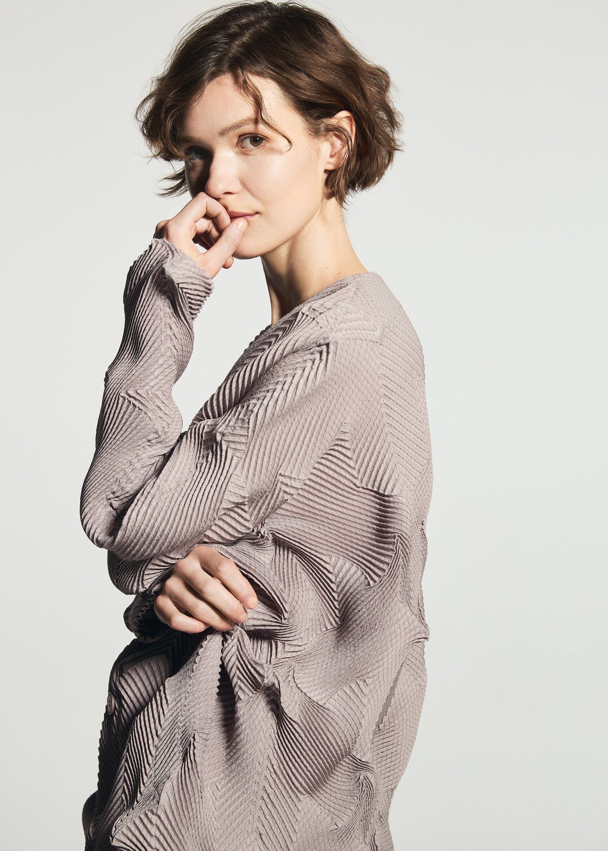 Zoomed view A person with short, wavy brown hair wears the TYPE-O 010-1 pleated cardigan by A-POC ABLE ISSEY MIYAKE, standing against a plain background and gazing thoughtfully at the camera with one hand near their face.