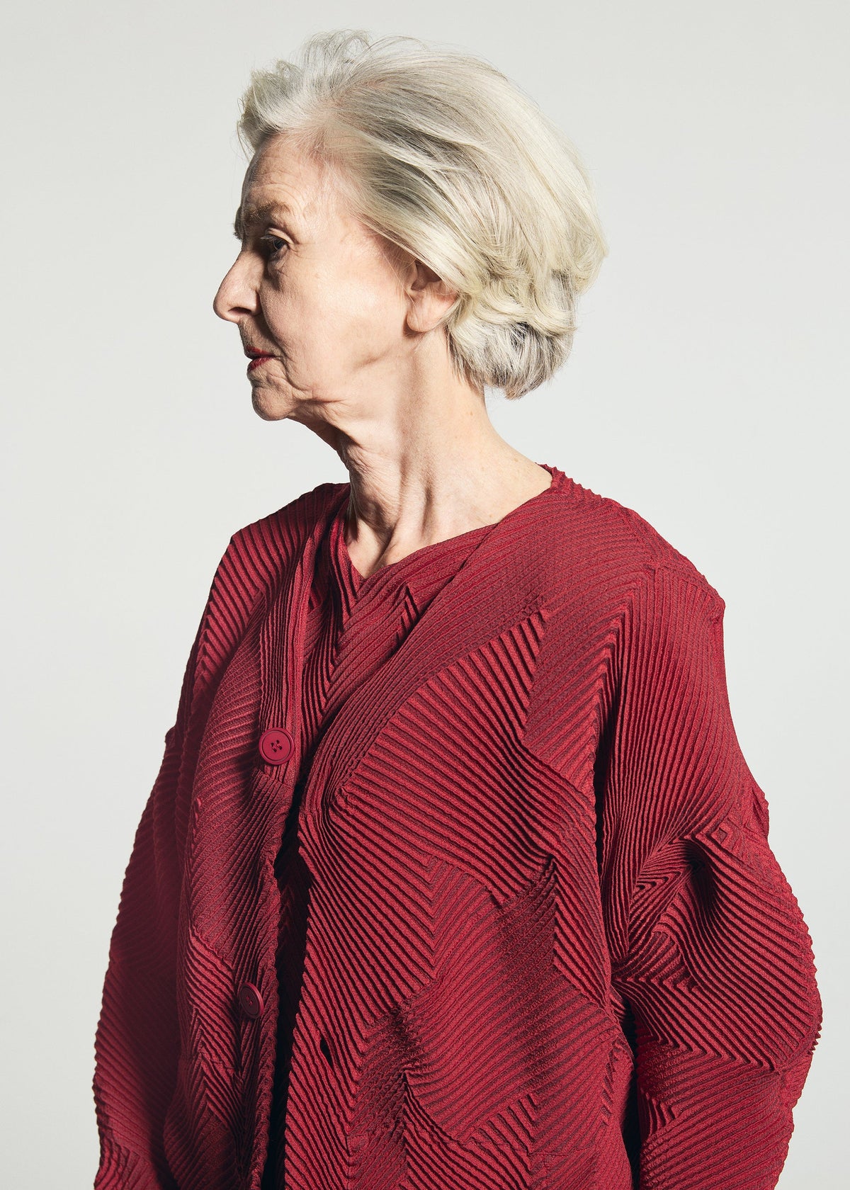 An elderly woman with short gray hair wears the TYPE-O 010-1 by A-POC ABLE ISSEY MIYAKE, a pleated red recycled polyester cardigan, standing in profile against a plain light background. view 9 of 13