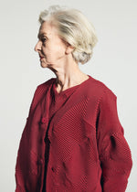 An elderly woman with short gray hair wears the TYPE-O 010-1 by A-POC ABLE ISSEY MIYAKE, a pleated red recycled polyester cardigan, standing in profile against a plain light background.