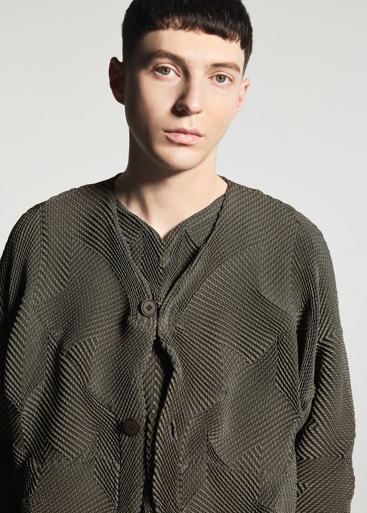 Zoomed view A person with short dark hair wears the TYPE-O 010-1 jacket by A-POC ABLE ISSEY MIYAKE, featuring textured olive-green fabric and large buttons, standing against a plain light grey background.