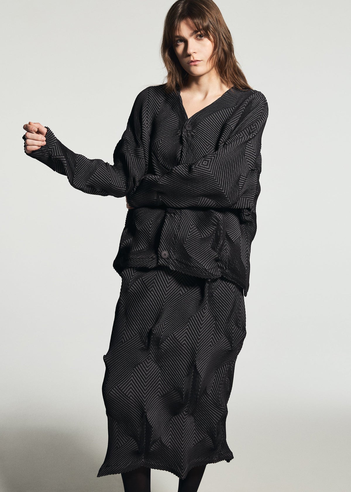 A woman poses in a relaxed stance wearing the TYPE-O 010-1 by A-POC ABLE ISSEY MIYAKE—a loose, houndstooth-patterned black jacket and matching skirt—against a plain, light-colored background. view 11 of 13