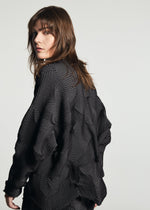 A person with shoulder-length brown hair wears the TYPE-O 010-1 by A-POC ABLE ISSEY MIYAKE, a black pleated cardigan with a geometric pattern, looking over their shoulder against a simple light background.