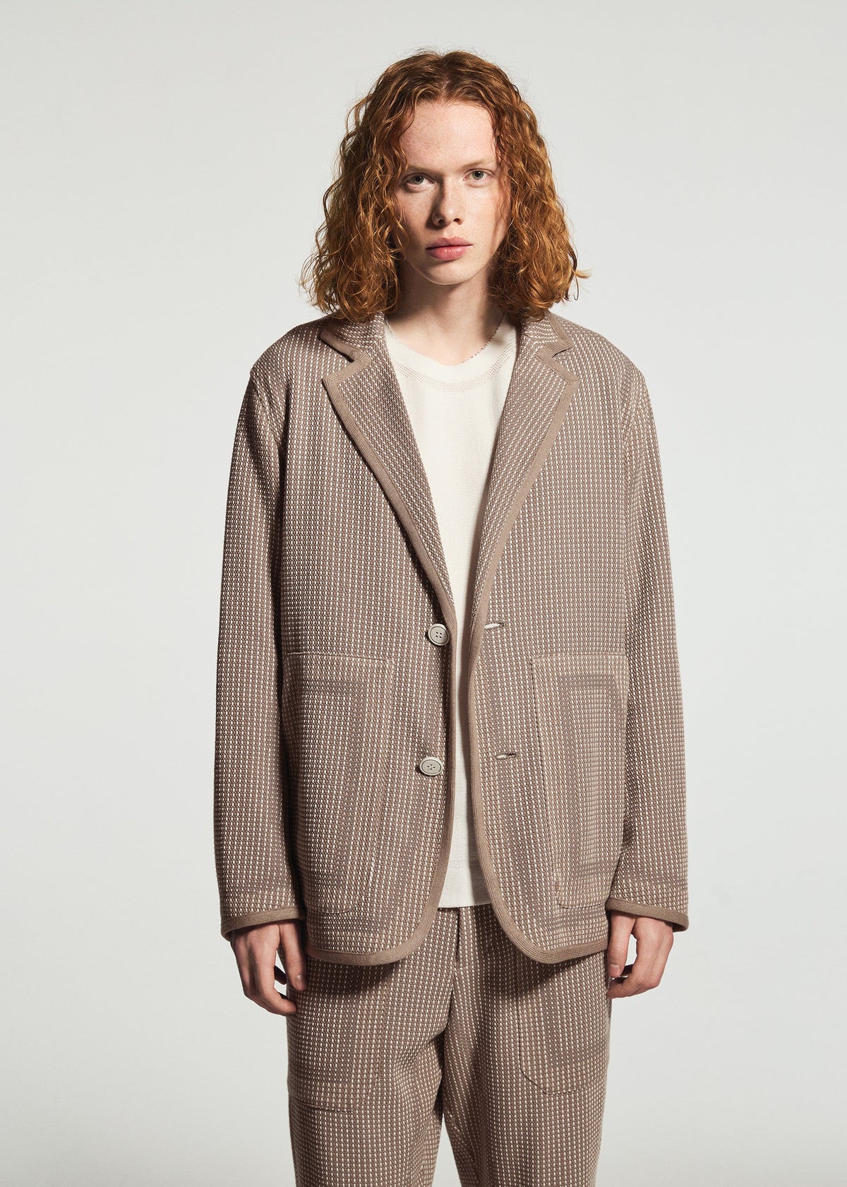 A person with shoulder-length curly red hair wears the TYPE-K 012 by A-POC ABLE ISSEY MIYAKE, a beige sashiko style suit jacket over a white shirt, standing against a plain white background. view 7 of 11