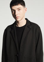 A person with short dark hair wears the TYPE-K 012 by A-POC ABLE ISSEY MIYAKE—a black textured sashiko-style jacket—over a black shirt, standing against a plain light background and looking slightly to the side.