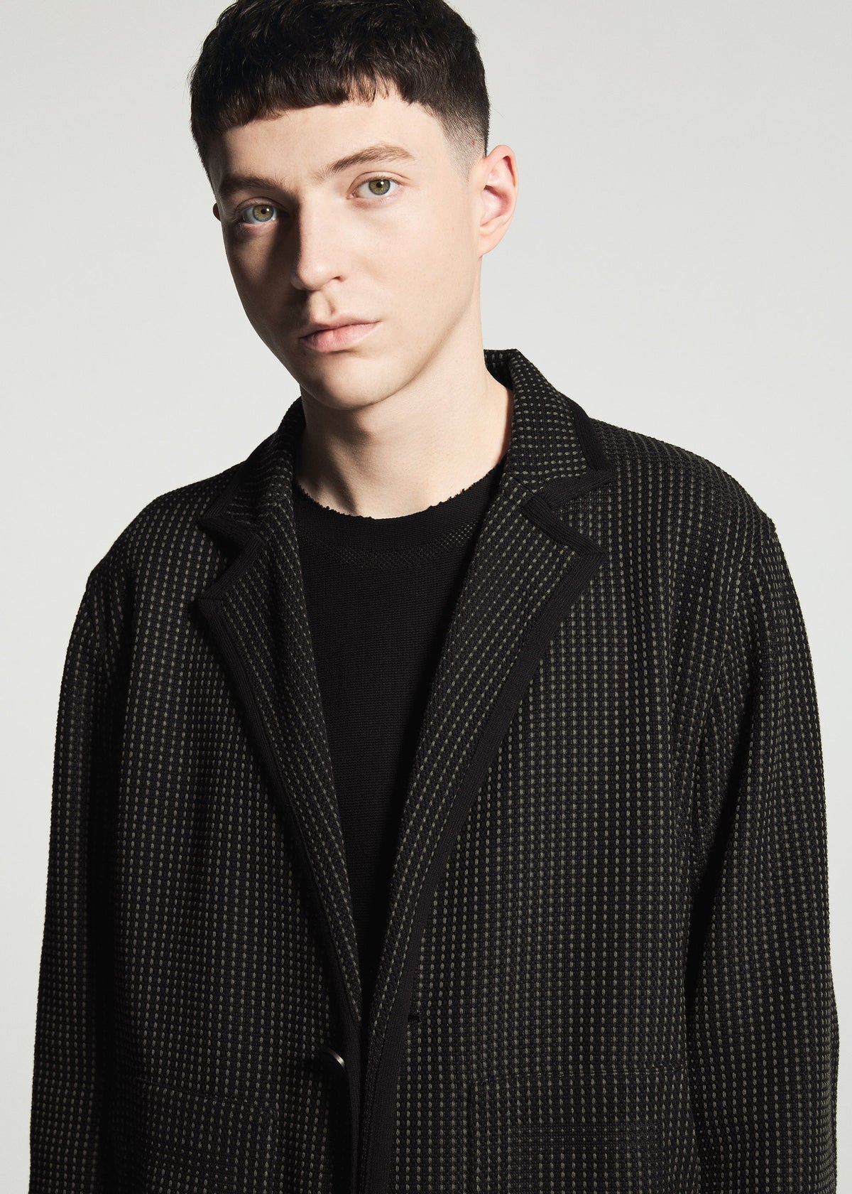 Zoomed view A person with short dark hair wears the TYPE-K 012 by A-POC ABLE ISSEY MIYAKE—a black textured sashiko-style jacket—over a black shirt, standing against a plain light background and looking slightly to the side.
