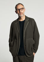 A man with short hair and glasses wears the TYPE-K 012 by A-POC ABLE ISSEY MIYAKE—sashiko style jacket and matching pants over a black shirt, standing against a plain, light background.
