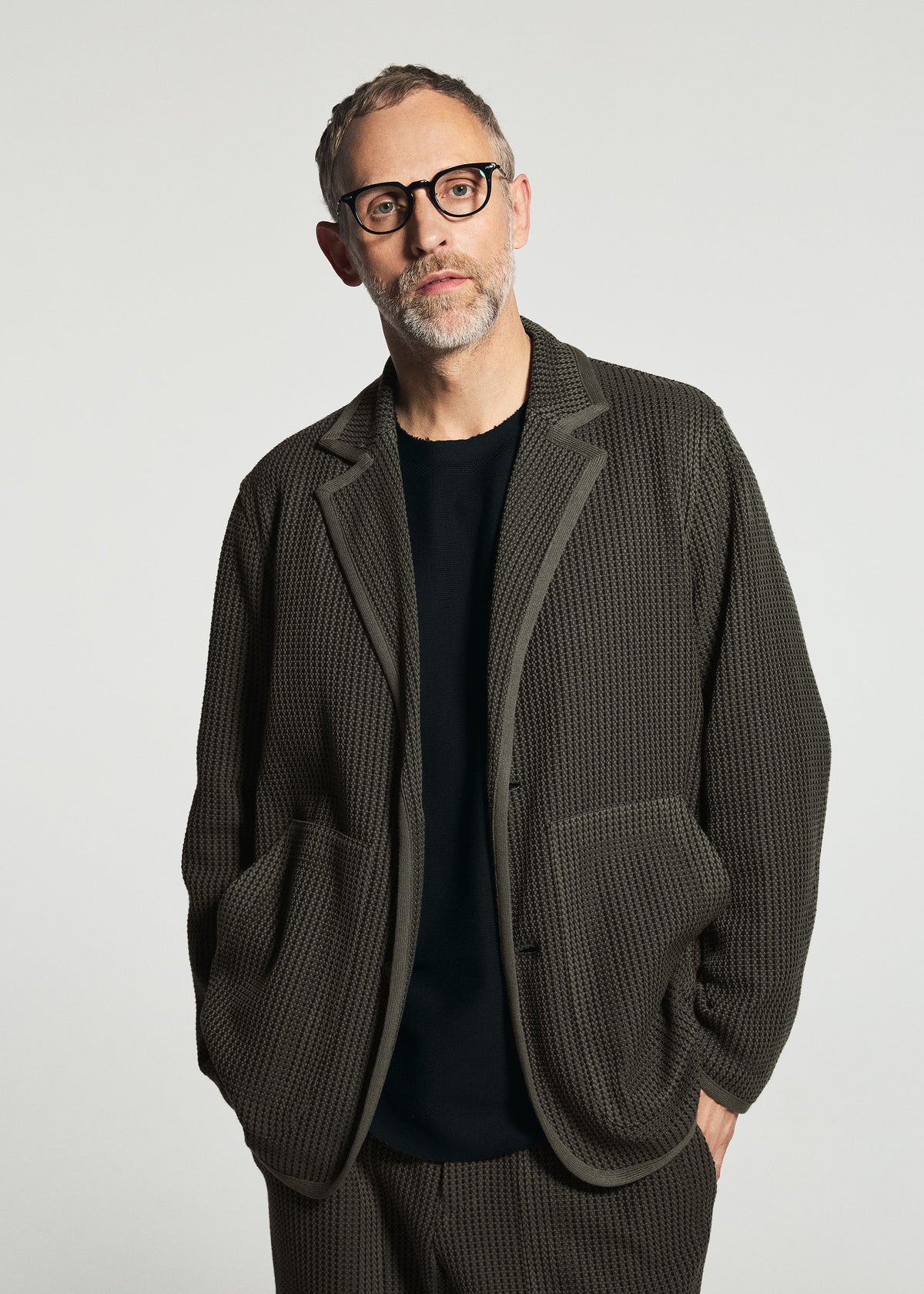 Zoomed view A man with short hair and glasses wears the TYPE-K 012 by A-POC ABLE ISSEY MIYAKE—sashiko style jacket and matching pants over a black shirt, standing against a plain, light background.