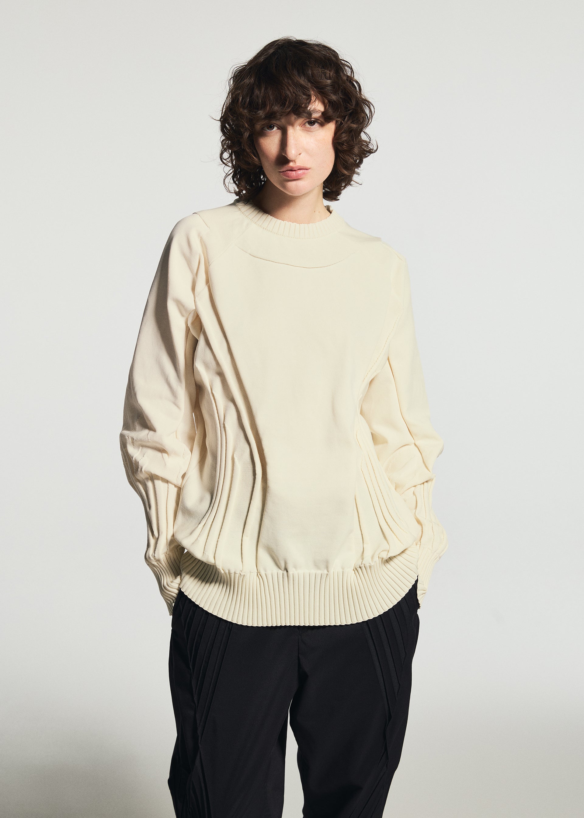 Zoomed view A person with short, curly brown hair wears the TYPE-K 001 by A-POC ABLE ISSEY MIYAKE—a cream-colored knit top featuring ribbed details and a distinctive Steam Stretch pattern crafted from stretchy polyester—paired with black pants. They stand against a plain light background, hands in pockets, while the unique textural design adds both comfort and style to the look.