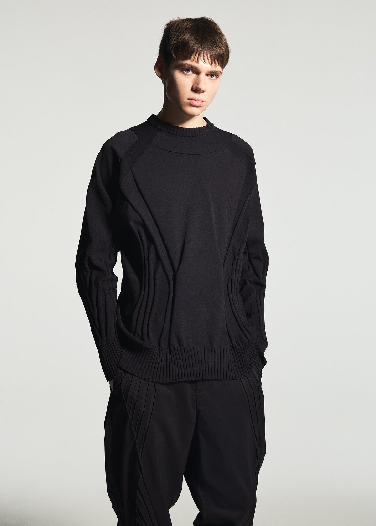 A young person with short brown hair wears the TYPE-K 001 by A-POC ABLE ISSEY MIYAKE—a textured black knit top featuring a Steam Stretch pattern, paired with matching stretchy polyester black pants—standing against a plain light background with a neutral expression. view 8 of 11