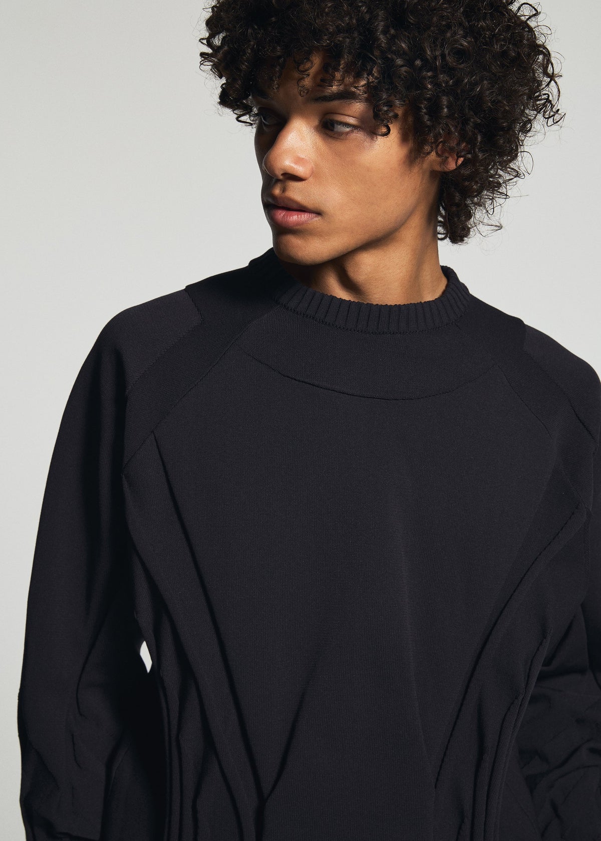 A person with curly hair, dressed in the TYPE-K 001 black stretchy polyester knit top by A-POC ABLE ISSEY MIYAKE, looks to their left against a plain light background. view 9 of 11