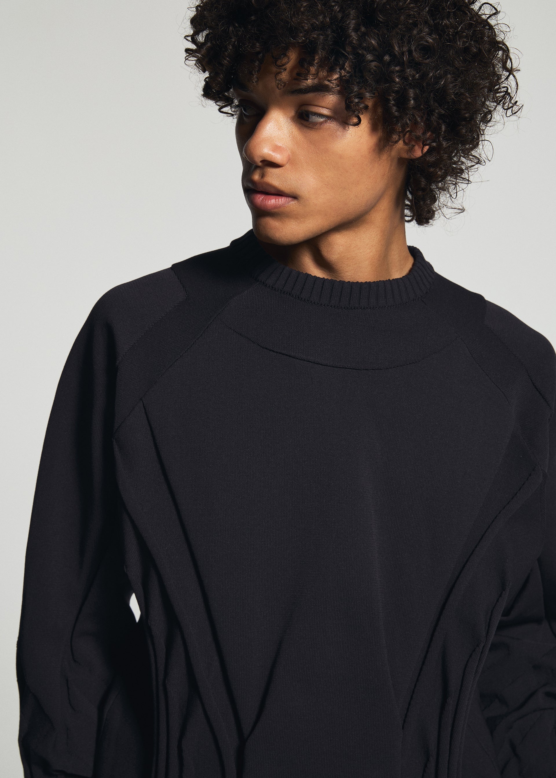 Zoomed view A person with curly hair, dressed in the TYPE-K 001 black stretchy polyester knit top by A-POC ABLE ISSEY MIYAKE, looks to their left against a plain light background.