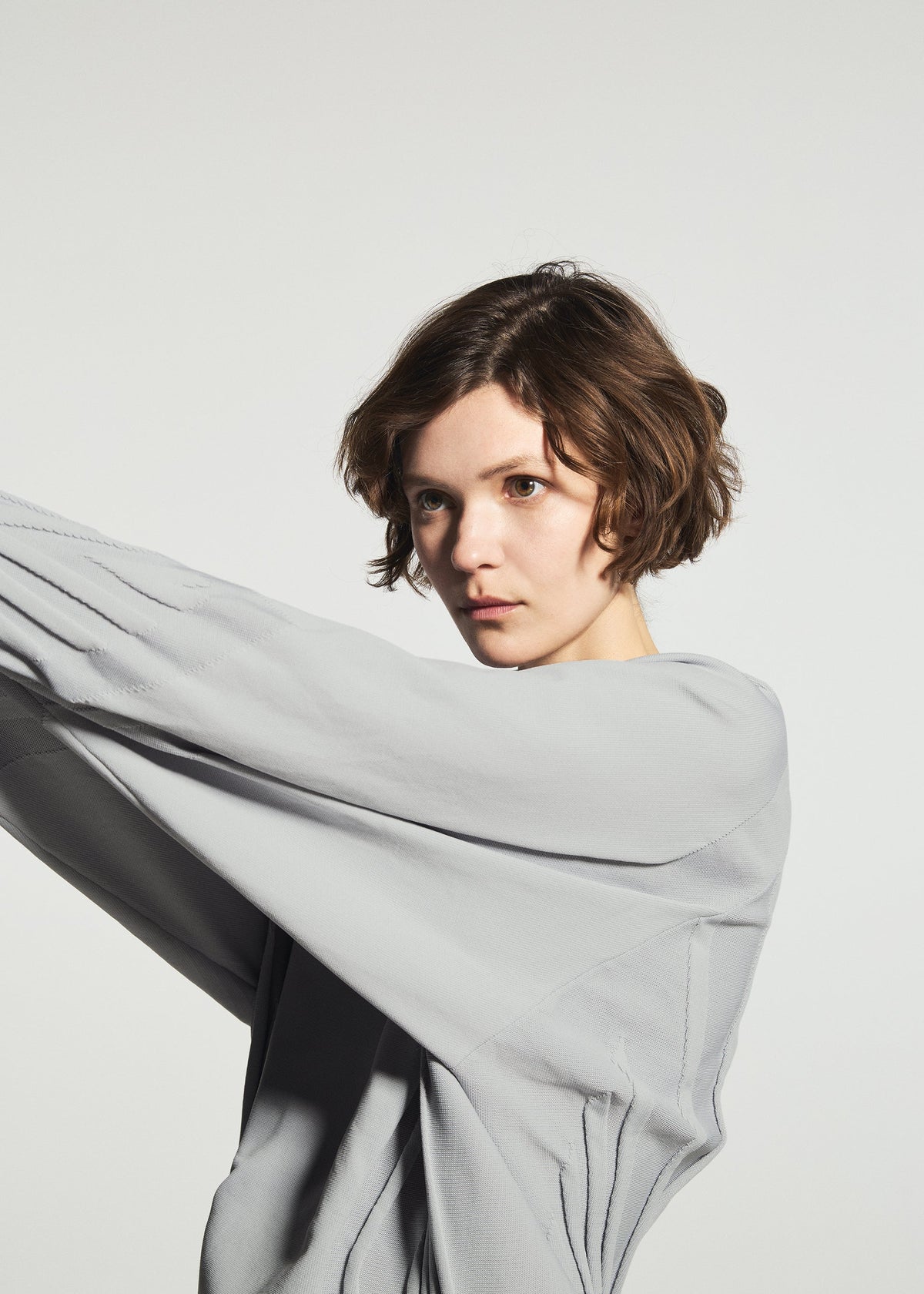 A person with short brown hair wears the A-POC ABLE ISSEY MIYAKE TYPE-K 001, a loose light gray knit top made from stretchy polyester with a Steam Stretch pattern, and poses with their arms raised to the side against a plain white background. view 10 of 11