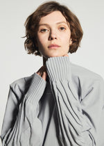 Wearing the TYPE-K 001 knit top by A-POC ABLE ISSEY MIYAKE, a person with short brown hair showcases the light gray garment’s ribbed cuffs and subtle Steam Stretch pattern, holding the sleeves up to their chin while looking directly at the camera against a plain background.