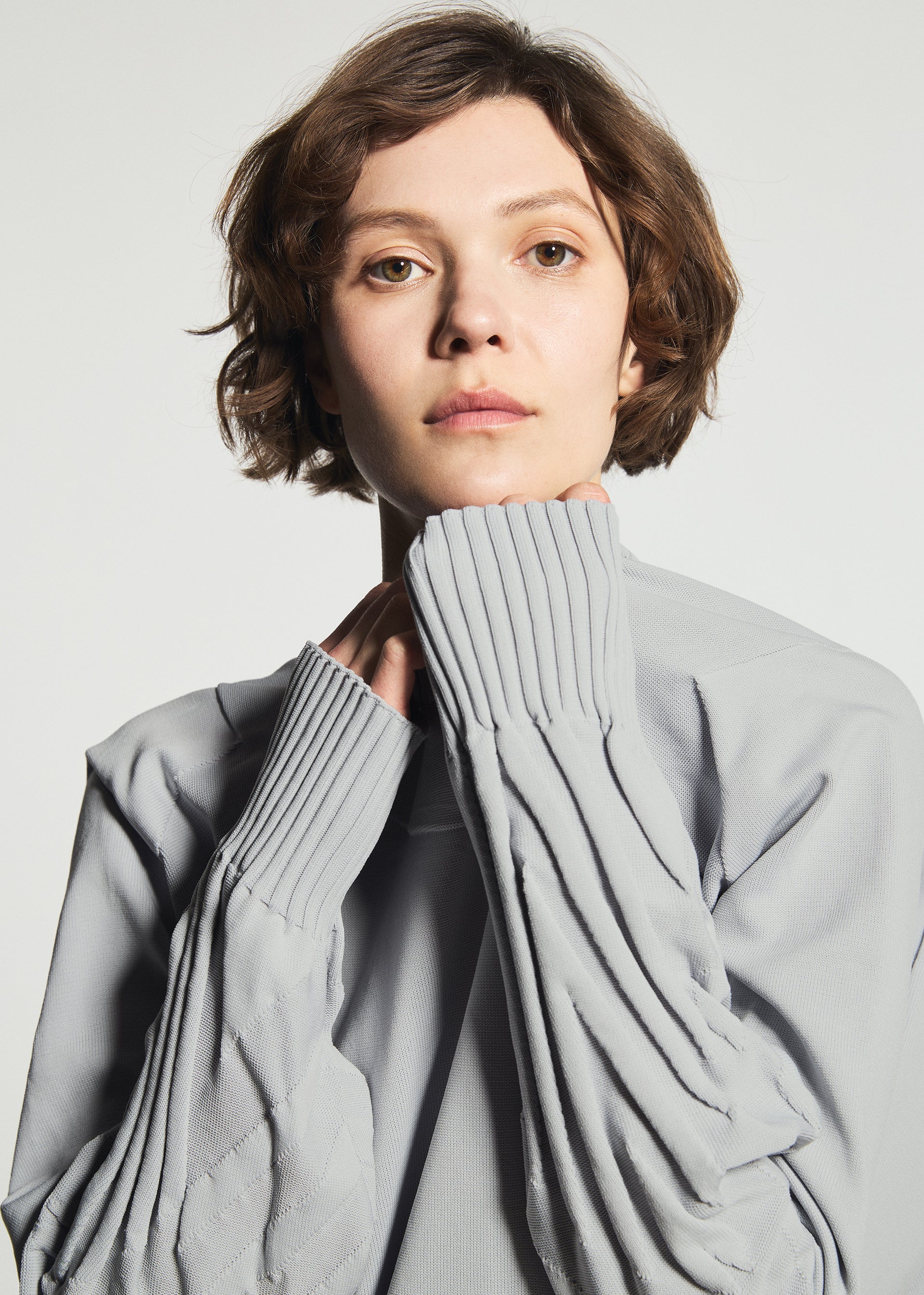 Zoomed view Wearing the TYPE-K 001 knit top by A-POC ABLE ISSEY MIYAKE, a person with short brown hair showcases the light gray garment’s ribbed cuffs and subtle Steam Stretch pattern, holding the sleeves up to their chin while looking directly at the camera against a plain background.