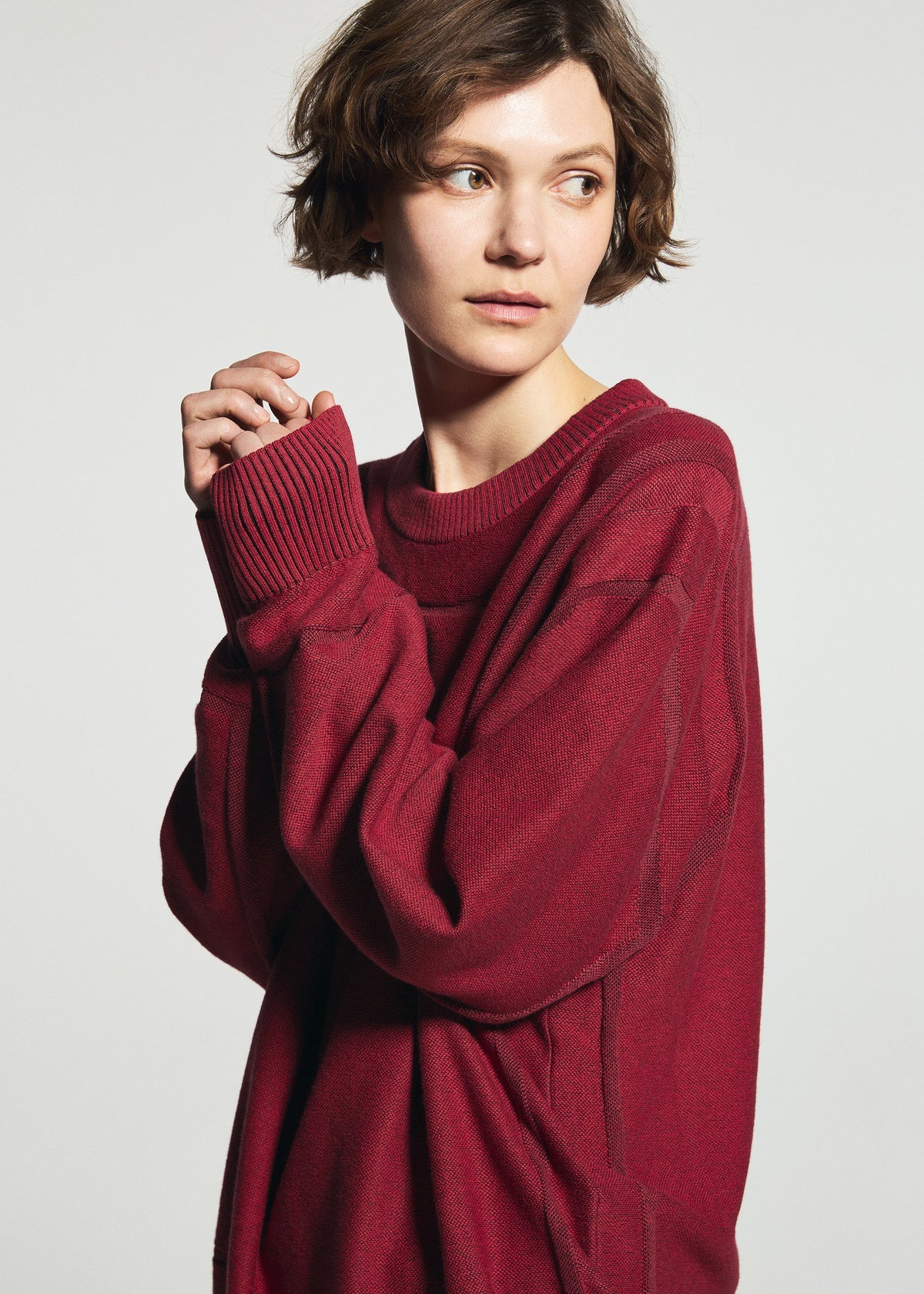 A person with short brown hair wears the TYPE-K 010 by A-POC ABLE ISSEY MIYAKE, gazing sideways with hands together near their face against a plain light background. view 7 of 8