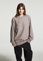 A person with straight brown hair wears the TYPE-K 010 by A-POC ABLE ISSEY MIYAKE, a loose taupe long-sleeved knit top, paired with black pants against a plain light background.