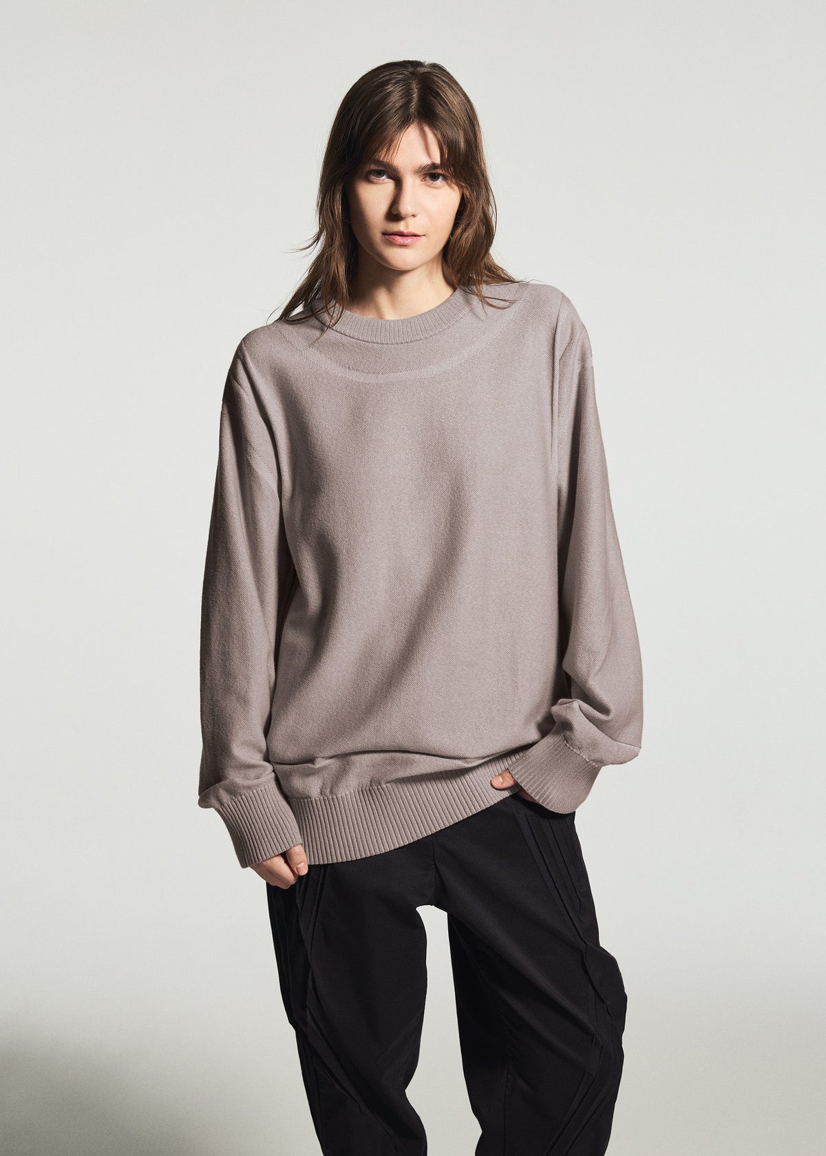 Zoomed view A person with straight brown hair wears the TYPE-K 010 by A-POC ABLE ISSEY MIYAKE, a loose taupe long-sleeved knit top, paired with black pants against a plain light background.